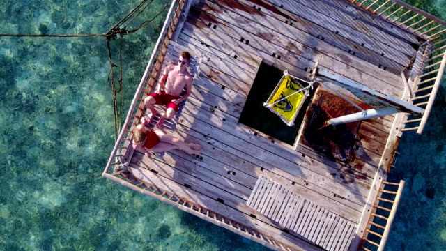 v03823-fliegenden-Drohne-Luftaufnahme-der-Malediven-weißen-Sandstrand-2-Personen-junges-Paar-Mann-Frau-entspannend-auf-sonnigen-tropischen-Inselparadies-mit-Aqua-blau-Himmel-Meer-Wasser-Ozean-4k-schwimmenden-Ponton-Steg-Sonnenbaden-zusammen