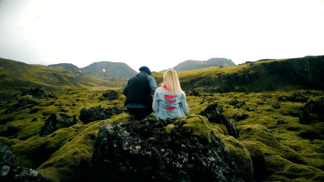 Vista-trasera-del-joven-hermosa-pareja-sentada-sobre-la-roca-y-disfrutando-el-paisaje-escénico-de-los-campos-de-lava-en-Islandia