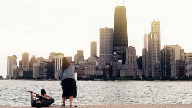 Young-creative-couple-on-the-shore-of-Michigan-lake,-Chicago,-America.-Woman-draw,-ma-play-guitar-lying-on-shore