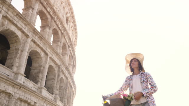 Beautiful-young-woman-in-colorful-fashion-dress-walking-alone-with-bike-arriving-in-front-of-colosseum-in-Rome-at-sunset-with-happy-attractive-tourist-girl-with-straw-hat-ground-shot