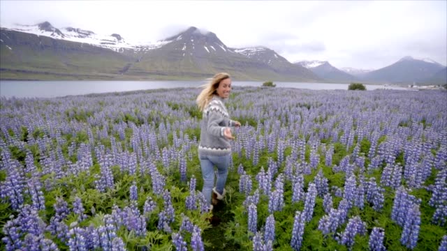 Follow-me-to-Iceland,-girlfriend-waving-hand-at-man-to-flower-lupine-field-near-lake-and-mountains-People-travel-concept--4K-video