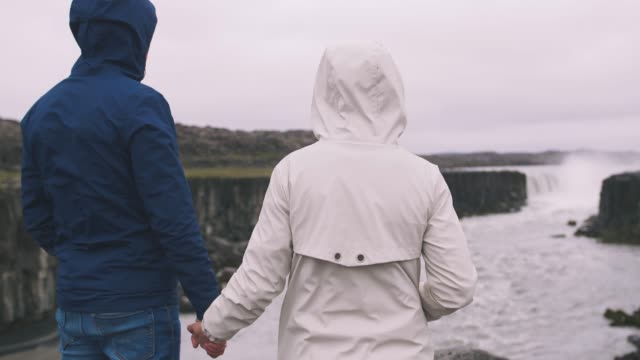 Back-view-of-young-hipster-couple-stranding-near-the-powerfull-waterfall-in-Iceland-and-enjoying-the-view