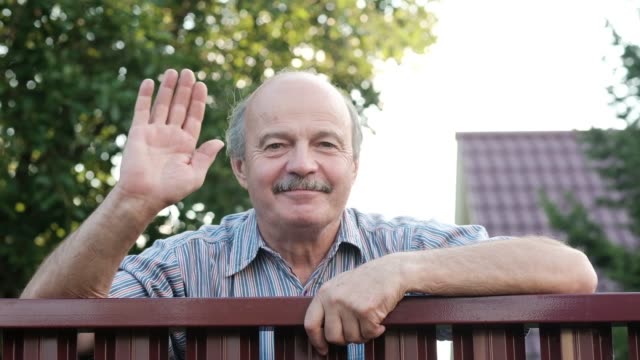 Friendly-caucasian-old-man-waving-hi-or-farewell,-isolated-outdoors-background-with-green-trees-and-fence