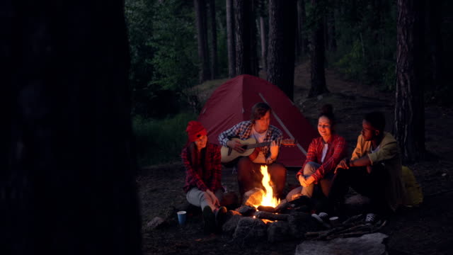 Multiracial-group-of-friends-hikers-is-playing-the-guitar,-singing-and-laughing-around-campfire-in-the-evening.-Summertime-fun,-nature-and-leisure-concept.
