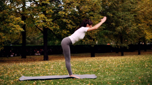 Side-view-of-slender-girl-is-sports-clothing-exercising-outdoors-doing-yoga-in-park-alone-on-warm-autumn-day.-Nature,-recreation-and-youth-concept.