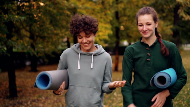 Beautiful-girl-is-walking-in-park-with-yoga-instructor-holding-mats-talking-gesturing-and-laughing.-Teacher-student-relationship-and-conversation-concept.