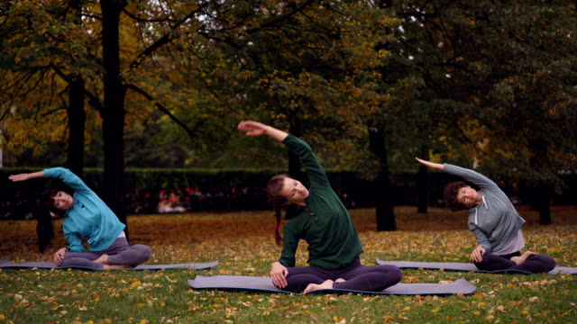 Instructora-de-yoga-de-mujer-es-mostrando-ejercicios-de-estiramiento-y-relajación-en-posición-de-loto-con-los-ojos-cerrados,-sentados-en-esteras-en-las-prácticas-al-aire-libre-en-el-parque.