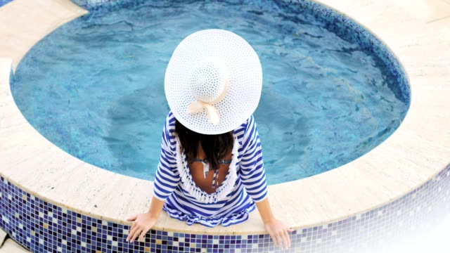 High-angle-rear-view-elegance-woman-in-hat-with-bow-sitting-on-edge-of-swimming-pool-or-hot-tub