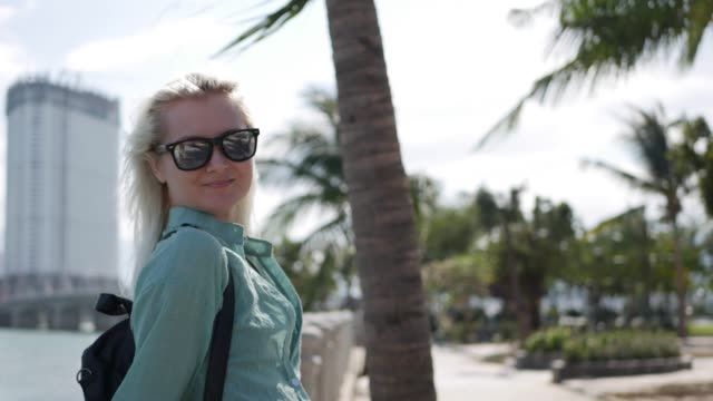 Young-happy-caucasian-woman-with-long-blonde-hair-in-sunglasses-and-green-shirt-standing-and-smiling-near-palm-tree-on-a-park-and-sea-background.-Travel-concept