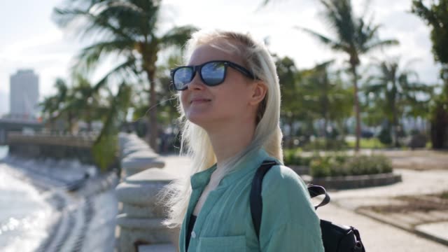 Glücklich-kaukasische-Mädchen-mit-langen-blonden-Haaren-mit-Sonnenbrille-und-grünes-Hemd-stehen-und-in-der-Nähe-von-Palme-auf-einen-Park-und-Meer-Hintergrund-lächelt.-Reisekonzept