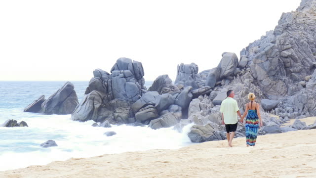 Eine-ältere-paar-Hände-halten,-zu-Fuß-am-Strand-entlang-in-vor-großen-Felsen