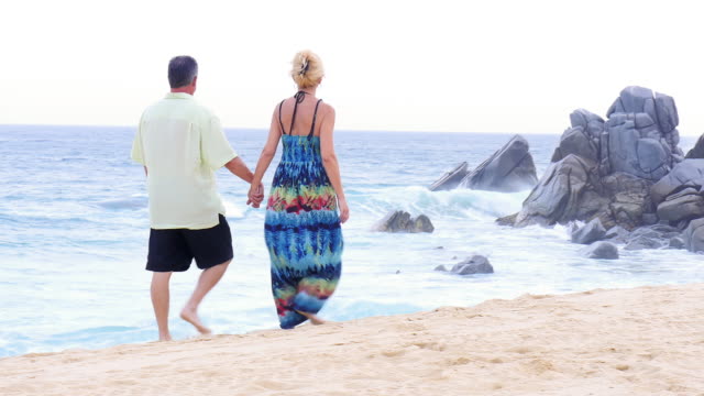 Una-Pareja-de-ancianos-sosteniendo-las-manos-y-caminar-en-la-playa,-en-frente-de-gran-rocas