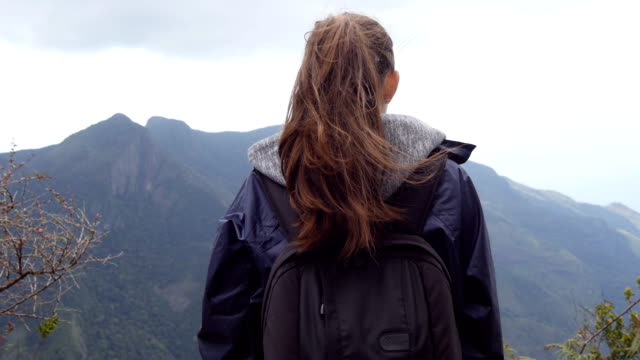 Unrecognizable-woman-tourist-standing-on-the-edge-of-beautiful-canyon-and-enjoying-landscapes.-Young-female-hiker-in-raincoat-with-backpack-reaching-up-top-of-mountain-and-raised-hands.-Rear-back-view