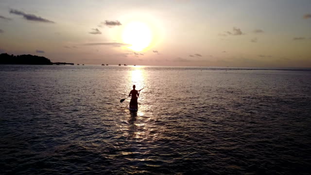 v04098-fliegenden-Drohne-Luftaufnahme-der-Malediven-weißen-Sandstrand-2-Personen-junges-Paar-Mann-Frau-Paddleboard-Rudern-Sonnenuntergang-Sonnenaufgang-auf-sonnigen-tropischen-Inselparadies-mit-Aqua-blau-Himmel-Meer-Wasser-Ozean-4k