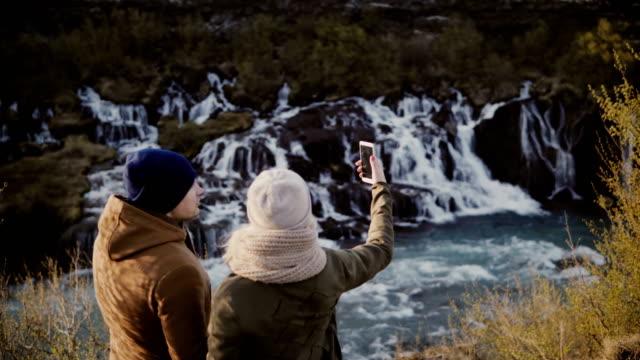 Young-traveling-couple-standing-in-mountains-valley-near-waterfall-and-talking-pictures-or-selfie-photos-on-smartphone