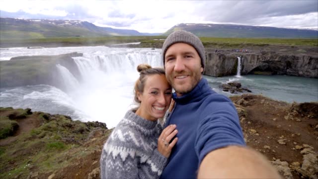 Travel-couple-fun-taking-selfie-photo-by-Godafoss-waterfall-on-Iceland-using-smartphone.-People-visiting-famous-tourist-attractions-and-landmarks-on-Route-1