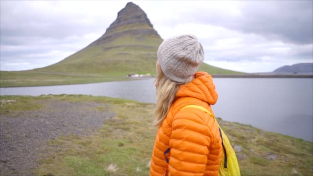 Mujer-joven-en-Islandia-contemplando-la-famosa-montaña-de-Kirkjufell,-reflexión-sobre-fiordo