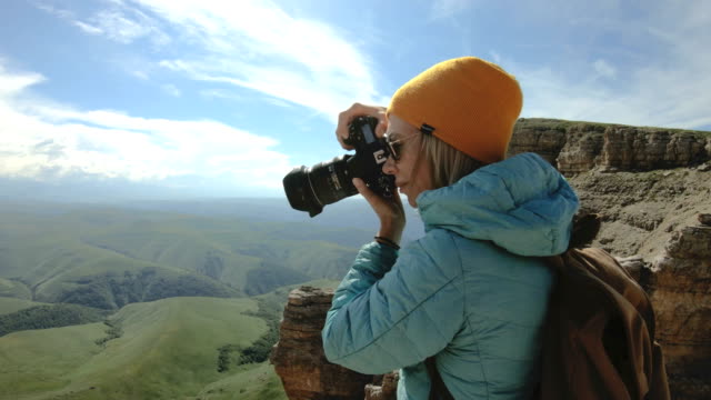 Blonde-girl-photographer-in-the-cap-takes-a-photo-on-her-digital-camera-with-a-background-of-rocks-in-the-Caucasus