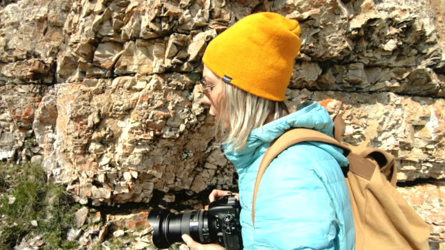 Blonde-girl-photographer-in-the-cap-takes-a-photo-on-her-digital-camera-with-a-background-of-rocks-in-the-Caucasus