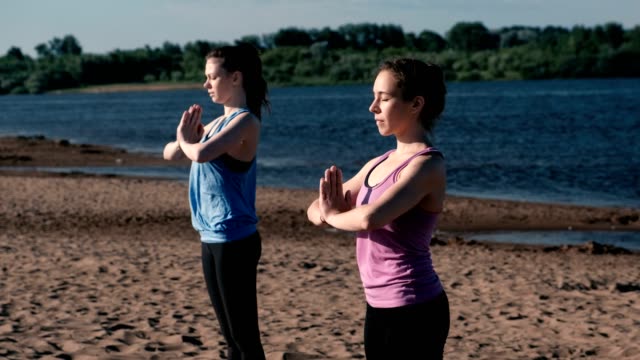 Two-woman-stretching-yoga-standing-on-the-beach-by-the-river-in-the-city.-Beautiful-city-view.-Namaste-pose.