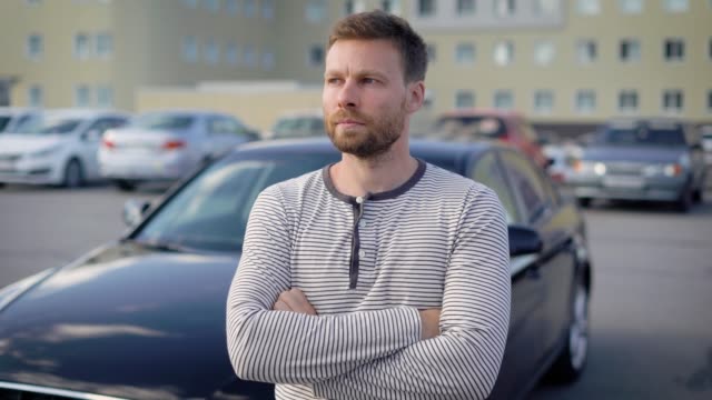 pensive-serious-man-is-standing-on-open-parking-area-in-summer-day,-crossed-hands-on-chest-and-looking-in-distance