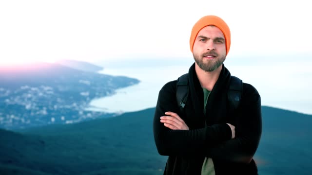 Portrait-European-friendly-hipster-man-standing-on-mountain-top-at-sunset