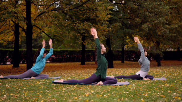 Group-of-young-ladies-is-doing-yoga-practising-King-Pigeon-pose-Eka-Pada-Rajakapotasana-on-mats-on-beautiful-green-and-yellow-lawn-in-park-on-autumn-day.
