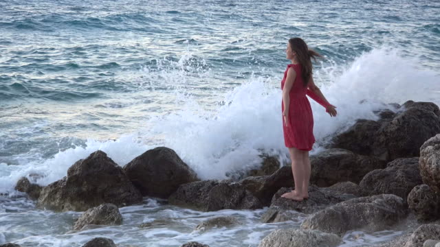 Beautiful-young-woman-on-rocky-seashore-open-arms-in-the-wind,-waves-splash