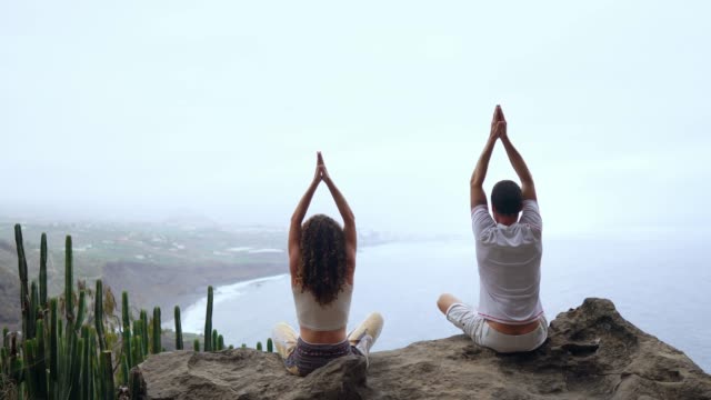 Ein-Mann-und-eine-Frau-sitzen-auf-einem-Berg,-der-Blick-auf-den-Ozean-sitzt-auf-einem-Stein-hob-ihre-Hände-und-die-Durchführung-eines-entspannenden-Atems-zu-meditieren.-Kanarischen-Inseln.