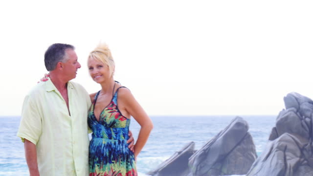 An-older-couple-smile-and-kiss-on-the-beach-with-waves-and-rocks-behind-them