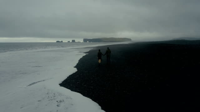 Luftaufnahme-des-Ehepaars-Hipster-zusammen-spazieren-am-Ufer-des-Meeres,-in-schwarzen-Vulkanstrand-in-Island