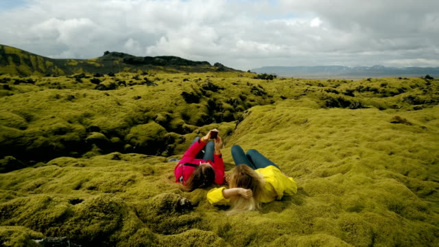 Vista-aérea-de-las-dos-mujeres-en-el-campo-de-lava-suave-en-Islandia.-Turistas-toman-selfie-en-el-smartphone