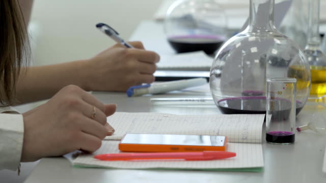 Los-estudiantes-en-el-trabajo-en-el-laboratorio-de-química-toman-notas-en-un-cuaderno.-Mujer-alumno-usando-el-teléfono-en-lección-de-ciencia.-Escritura-de-jóvenes-estudiantes-de-medicina