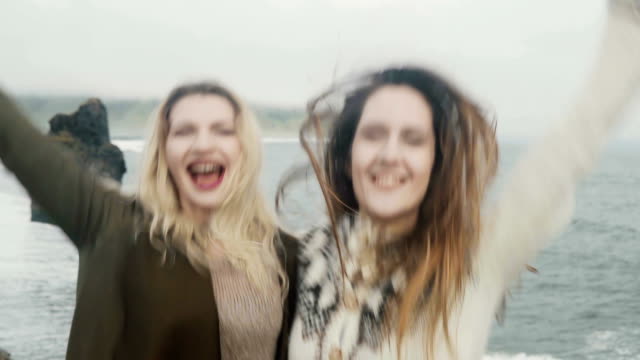 Retrato-de-mujer-feliz-dos-de-pie-en-la-playa-negra-en-Islandia-y-divertirse-juntos,-saltando-y-sonriendo