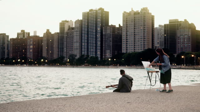 Young-Beautiful-couple-on-the-shore-of-Michigan-lake,-Chicago,-America.-Woman-draw,-man-play-guitar