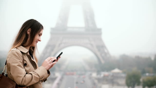 Young-beautiful-woman-with-mobile-phone-in-foggy-morning.-Female-using-smartphone-near-the-Eiffel-tower-in-Paris,-France