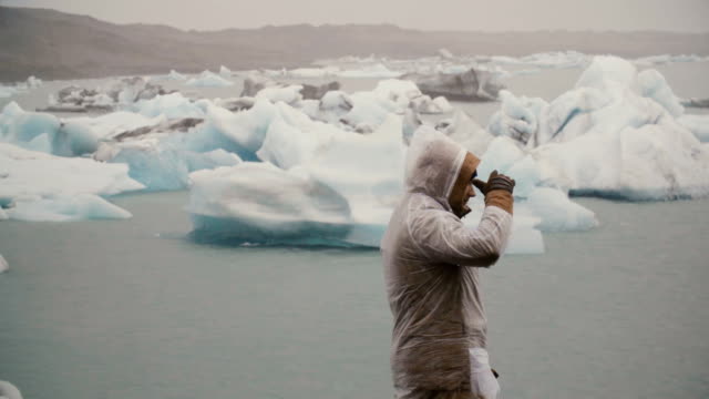 Back-view-of-young-man-in-raincoat-standing-in-ice-lagoon-in-Iceland.-Tourist-exploring-the-famous-sight-alone