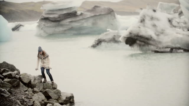 Young-happy-woman-walking-on-the-shore-of-ice-lagoon.-Tourist-in-lopapeysa-exploring-the-sights-of-Iceland