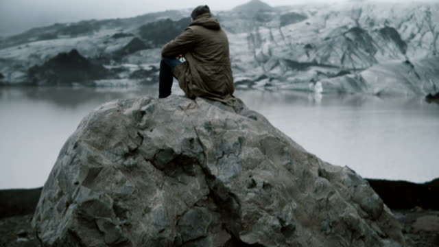Vista-trasera-del-hombre-joven-sentado-en-la-roca-volcánica-sola-y-mira-en-los-glaciares-de-la-laguna-de-hielo-en-Islandia
