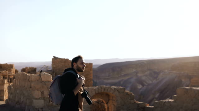 Professional-photographer-explores-ancient-ruins.-European-man-with-camera-and-backpack-near-mountain-view.-Israel-4K