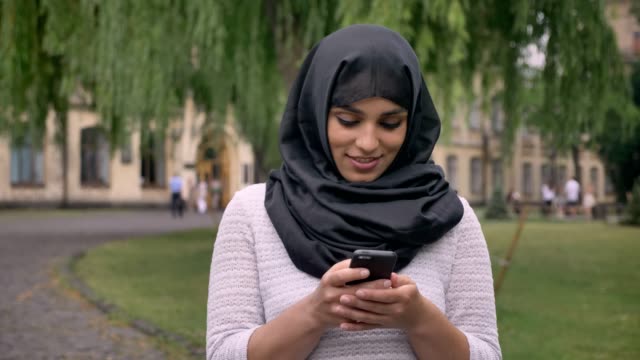 Young-beautiful-muslim-girl-in-hijab-is-typing-message-on-her-smartphone,-watching-at-camera-in-blue-weather,-communication-concept,-religious-concept