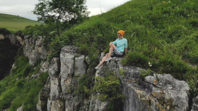 Young-hipster-woman-is-sitting-on-the-edge-on-the-grass-of-tall-wall-of-rock.-Aerial-view.-Drone-is-flying-slow-backward-from-model.-Establishing-revealing-shot