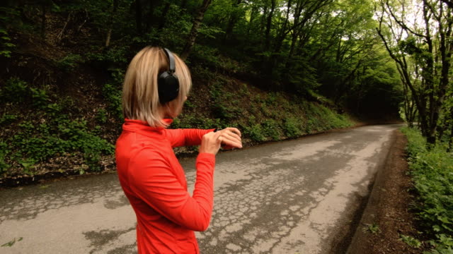 Junge-attraktive-Frau-im-Kopfhörer-Ändern-der-Einstellungen-auf-eine-Smartwatch-vor-oder-Joggen-auf-der-Straße-in-einem-grünen-Wald-psole