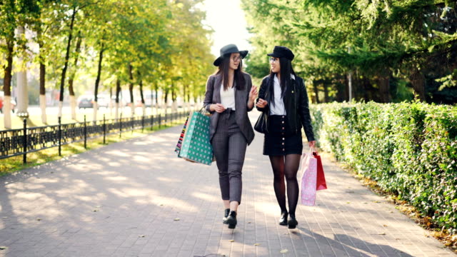 Dolly-shot-of-happy-girls-shopaholics-walking-in-the-street-carrying-shopping-paper-bags-and-talking-discussing-new-clothes-collection.-Youth,-leisure-and-consumerism-concept.