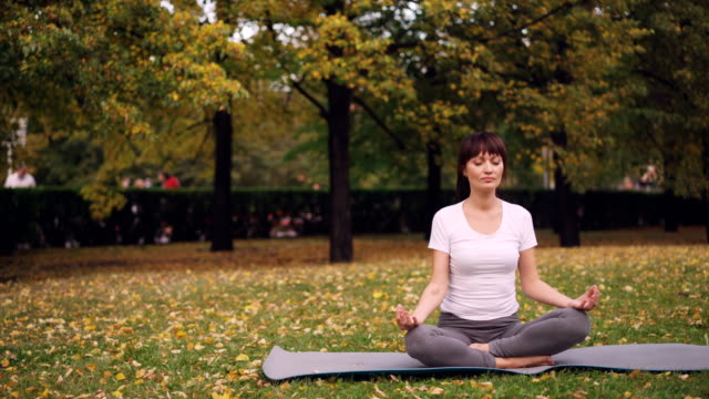 Attraktive-junge-Dame-sitzt-im-Lotussitz-auf-Yoga-Matte-im-Park,-die-Hand-in-Hand-in-Mudra-auf-Knien-und-Atmung-Entspannung-nach-dem-Training.-Meditation-und-Natur-Konzept.