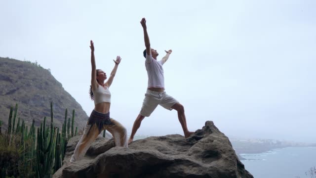 Un-hombre-y-una-mujer-de-pie-en-el-borde-de-un-acantilado-con-vistas-al-océano-levantan-sus-manos-y-aspirar-el-aire-del-mar-durante-yoga