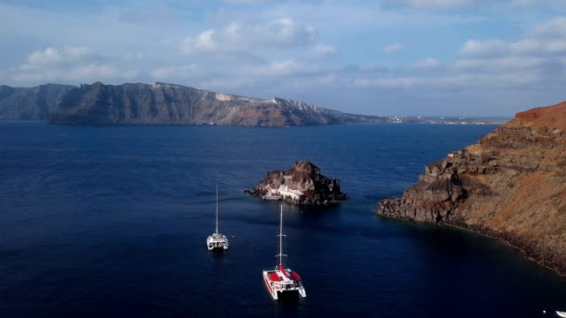 Flight-over-small-island-church-(Nisis-Agios-Nikolaos)-near-Oia-town,-Santorini-island,-Greece