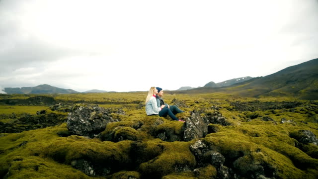 Vista-aérea-de-pareja-hermosa-joven-sentada-y-disfrutando-del-paisaje-del-campo-de-lava-en-Islandia.-Helicóptero-volando-alrededor