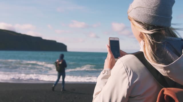 Young-loving-couple-taking-pictures-on-smartphone-on-beautiful-beach-in-Iceland-during-sunset,-slow-motion