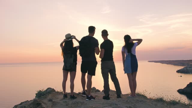 Gruppe-von-jungen-Wanderer-Freunden-reden,-erstaunliche-Meer-und-schönen-Sonnenuntergang-stehen-am-Felsen
